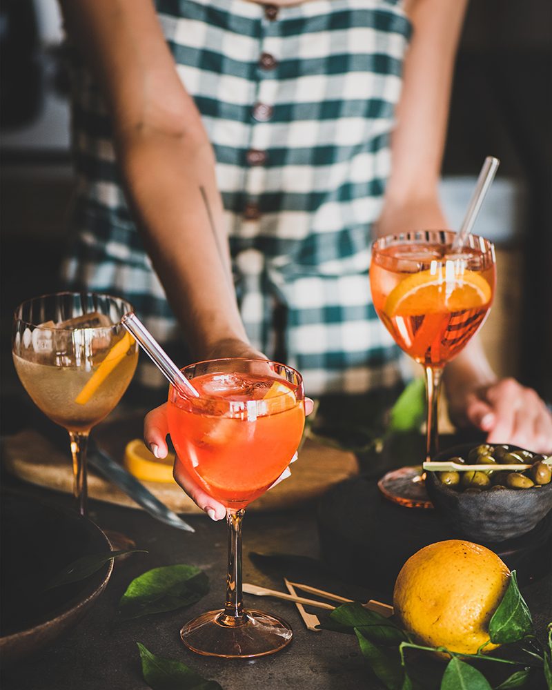 Young woman in checkered dress holding Aperol Spritz aperitif drink cocktail with orange in glass in kitchen. Summer refreshing drink concept