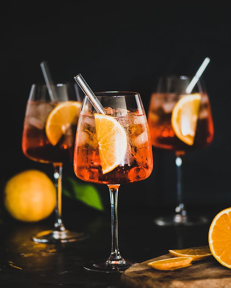 Aperol Spritz aperitif with oranges and ice in glass with eco-friendly glass straw on concrete table, black background, selective focus, copy space. Summer refreshing drink concept